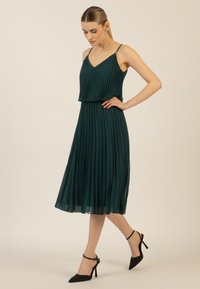 Grün gefälteltes Kleid mit V-Ausschnitt und verstellbaren Trägern, das ein geschichtetes Design aufweist. Getragen mit schwarzen High Heels.