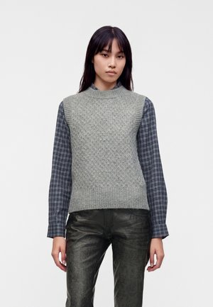 Jeune femme portant un pull sans manches gris en tricot par-dessus une chemise à carreaux bleue et un pantalon texturé foncé, debout devant un fond uni.