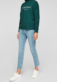Grüne Sweatshirt mit dem Text "conscious"; hellblaue Skinny-Jeans mit fransigem Saum; weiße Sneakers; lässiger Schnitt und bequemer Stoff.