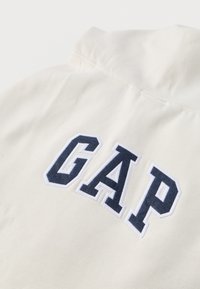 Valge puuvillane pullover, millel on esile tõstetud tumesinine "GAP" logo paksus kirjas. Omab lihtsat kraed ja siledat kangapinda.