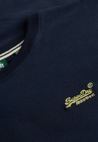 Marineblåt stof med broderet gult "SuperDry"-logo og japanske tegn, viser ribstrikket rundhalskrave med stribet indre detalje.