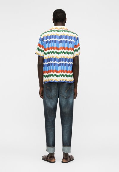 Άνδρας που στέκεται με την πλάτη γυρισμένη, φοράει πολύχρωμο πουκάμισο με ζιγκ ζαγκ μοτίβο, σκουρόχρωμα τζιν με γυρισμένα μπατζάκια και καφέ σανδάλια.