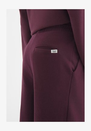 Burgundy joggers made of soft fabric, featuring a side pocket and a small label with "Chelsea Peers" on the back.
