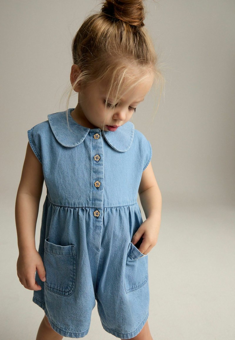 Combinaison en denim pour tout-petits de couleur bleu clair avec des manches courtes, un col arrondi, cinq boutons en bois et deux poches à l'avant. Texture douce, design décontracté.