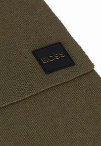 Bonnet en tricot vert olive avec un revers replié et une étiquette noire rectangulaire embossée portant le mot "BOSS" en lettres dorées.