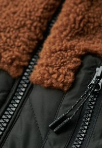 Veste isolante noire avec un col en fourrure brun, tissu texturé, fermeture à double zip et cordons de serrage noirs réglables.