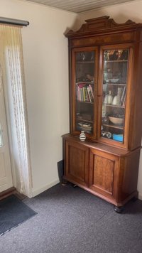 Armario de madera con un frente de vidrio, exhibiendo libros y artículos decorativos. Color marrón rico, detalles intrincados, tres estantes y patas redondas.