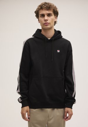 Jonge man draagt een zwarte hoodie met witte en rode gestreepte mouwen en beige broek, staand tegen een effen lichte achtergrond.
