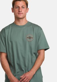 Grünes Baumwoll-T-Shirt mit rundem Ausschnitt und kurzen Ärmeln. Mit einem kreisförmigen Logo mit Berggrafiken und Text auf der linken Brust.