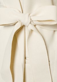Tissu doux de couleur crème noué en un grand nœud plat avec de larges boucles et de longs pans, mettant en avant la texture et les détails des coutures.
