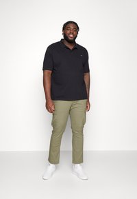 Čierne polo tričko a olivové cargo nohavice s prednými vreckami. Biele tenisky dotvárajú outfit. Jednoduché pozadie, neutrálny osvetlenie.