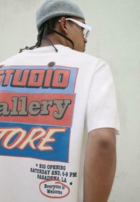 Bela majica s pisanjem grafičnim oblikom, rdečimi in modrimi blokov, krepko besedilo "STUDIO," "gallery," "STORE," in podrobnosti dogodka na dnu.