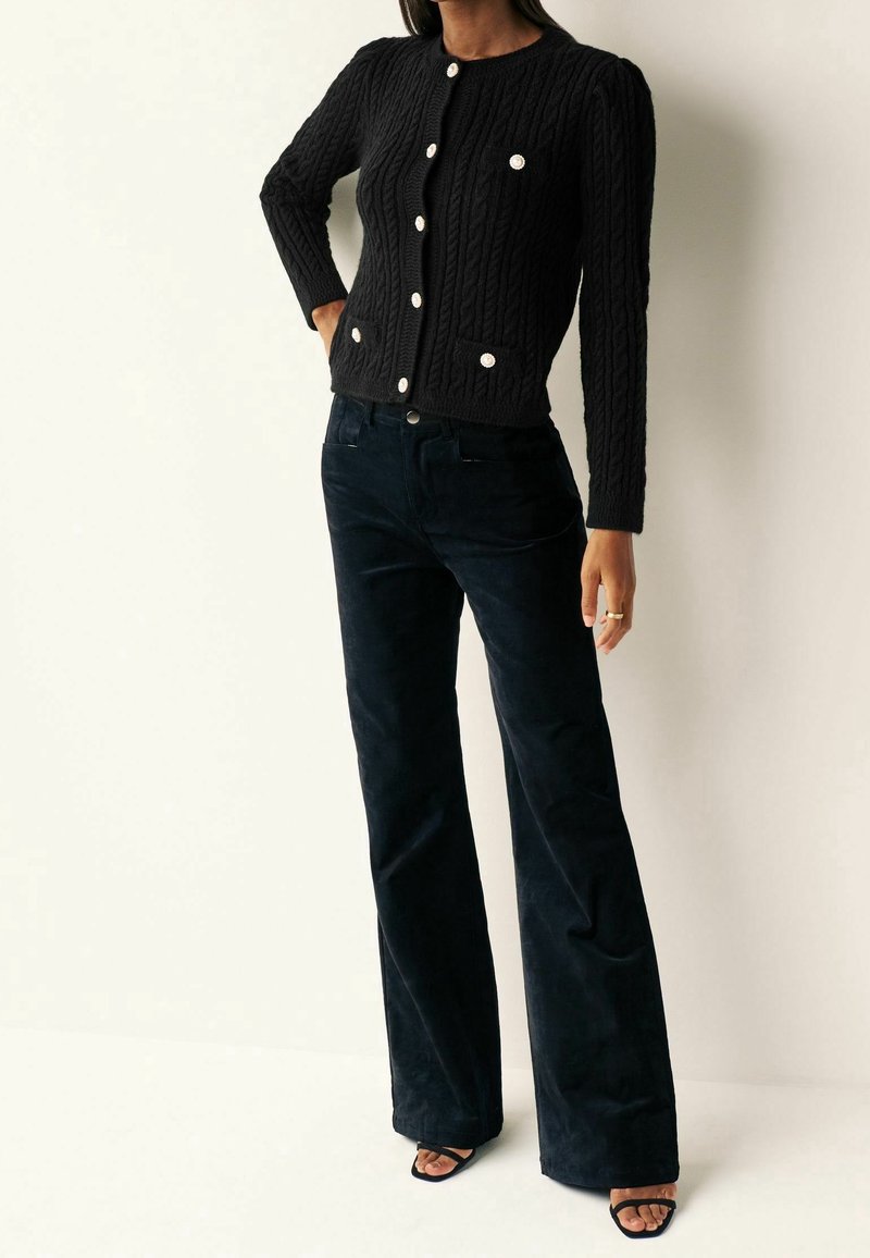 Czarny kardigan z plecionki z okrągłym dekoltem, z przodu na guziki, z dwoma kieszeniami; zestawiony z ciemnymi, rozkloszowanymi spodniami z kordurowej tkaniny i czarnymi sandałkami na obcasie.