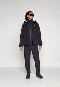 Black waterproof jacket with a hood, zippered front, and logo on chest, paired with matching pants and black boots. Smooth texture.