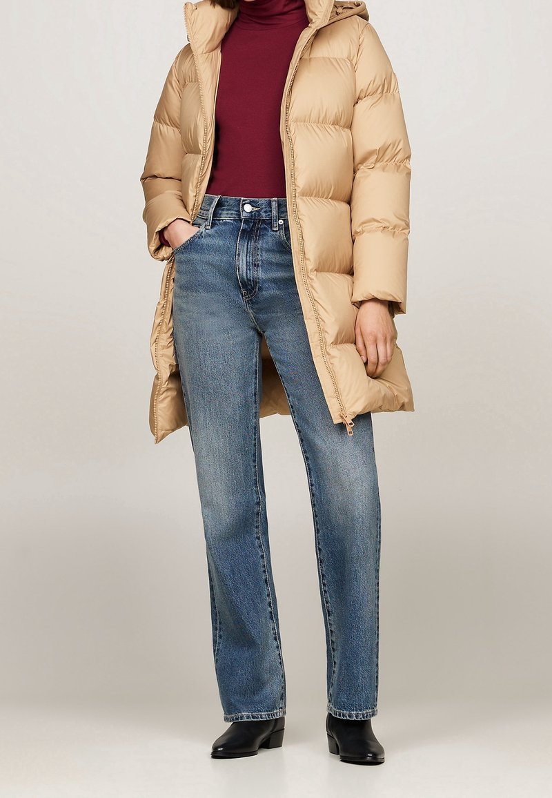 Beige pufferjack met capuchon, geribbelde maroon coltrui en blauwe rechte jeans. Zwarte enkellaarzen maken de outfit compleet.