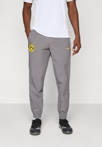 Homme portant un pantalon de sport gris avec le logo de Borussia Dortmund et l'emblème jaune de Puma, associé à des chaussures de course noires et des chaussettes blanches.