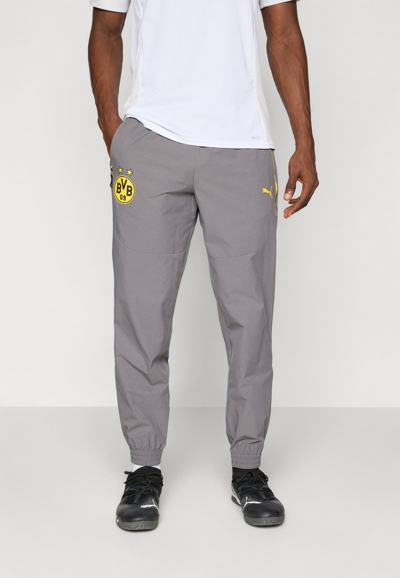 Homme portant un pantalon de sport gris avec le logo de Borussia Dortmund et l'emblème jaune de Puma, associé à des chaussures de course noires et des chaussettes blanches.