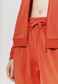 Mi-corps d'une personne portant une veste orange ouverte zippée et un pantalon assorti à taille élastique avec un cordon noué.
