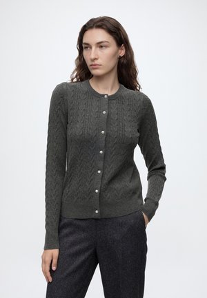Femme aux longs cheveux bruns portant un cardigan gris foncé à torsades avec des boutons en perle et un pantalon texturé foncé, debout, une main dans la poche.