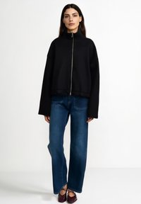 Črno pulover z zadrgo in visokim ovratnikom, kratki kroj in široki rokavi, kombiniran z modrimi širokimi kavbojkami in bordo čevlji.