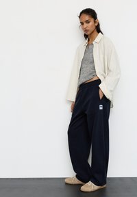 Młoda kobieta stoi przy białej ścianie, ubrana w luźną kremową koszulę, szary top, granatowe szerokie spodnie dresowe oraz beżowe buty sznurowane.