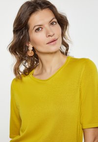 Kortærmet, rundhalset bluse i lys gul, lavet af struktureret stof. Matches med runde, ferskenfarvede øreringe med en guldfarvet detalje.