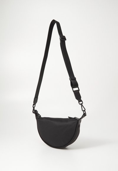 Must mustapeaga mustaristidega kott, valmistatud nailonist, millel on sile tekstuur, reguleeritav rihm ja lukuga sulgemine. Omab metallist detailide aktsente.