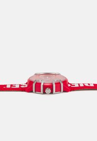 Red wristwatch with a round plastic case, transparent dial, and silver-tone accents. Features a textured red fabric strap with white lettering.