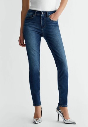 Vrouw draagt donkerblauwe skinny jeans, wit shirt en zilveren hoge hakken, met één hand in haar zak en de andere ontspannen langs haar zijde.