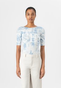 Top fitted a maniche corte in tie-dye blu con grande logo bianco "GUESS", abbinato a pantaloni chiari, tessuto morbido e materiale elasticizzato.