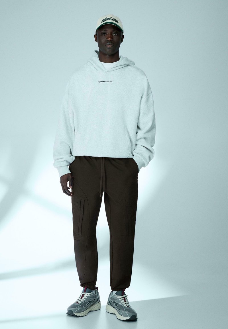 Homme portant un sweat à capuche gris clair, un pantalon cargo foncé, des baskets grises et une casquette blanche, debout devant un fond clair uni.