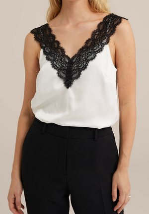 Vrouw draagt een witte mouwloze blouse met een zwarte kanten V-hals en zwarte hoge taille broek tegen een neutrale achtergrond.