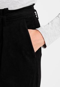 Černé semišové kalhoty se svázaným pásem, které mají boční kapsy a hladký povrch. Také je částečně vidět šedý top s dlouhým rukávem.