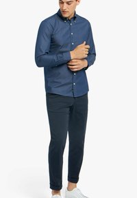 Marine knoop-overhemd, lange mouwen, katoenen stof, slim fit, gecombineerd met een donkere chino en witte sneakers, staand met één hand op de arm.