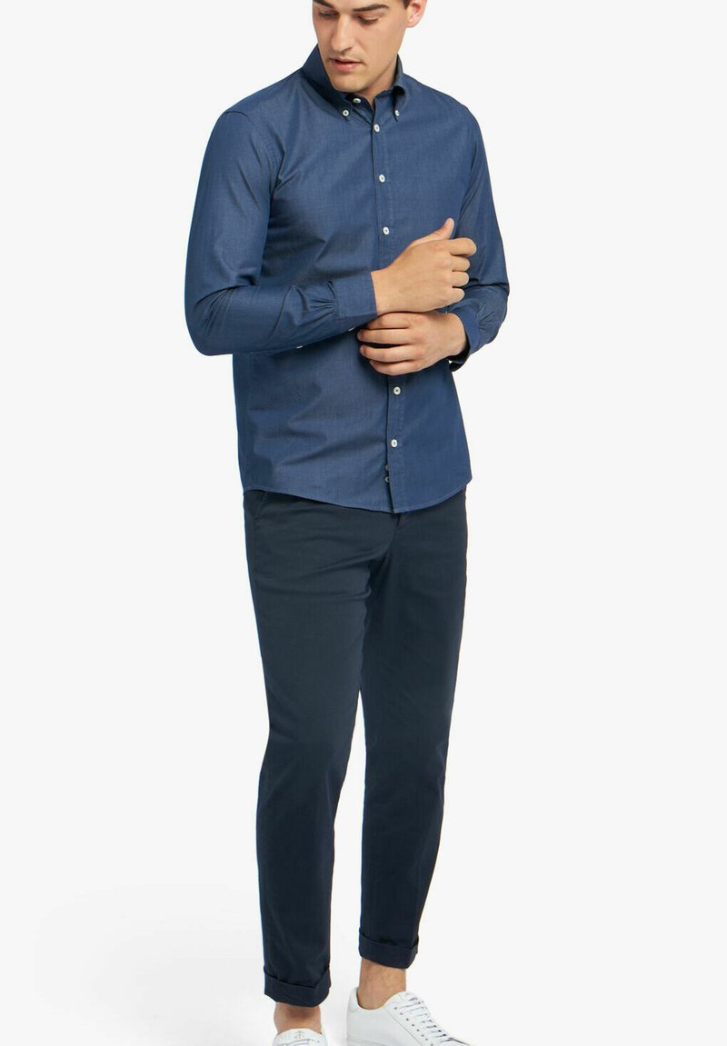 Marine knoop-overhemd, lange mouwen, katoenen stof, slim fit, gecombineerd met een donkere chino en witte sneakers, staand met één hand op de arm.