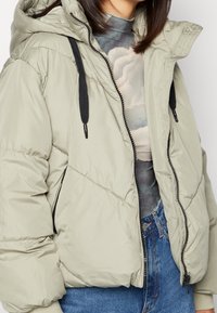 Gewatteerd, lichtgrijs jack met een capuchon; voorzien van zwarte trekkoorden en een ritssluiting. Draagt over een top met patroon en gecombineerd met een blauwe spijkerbroek.