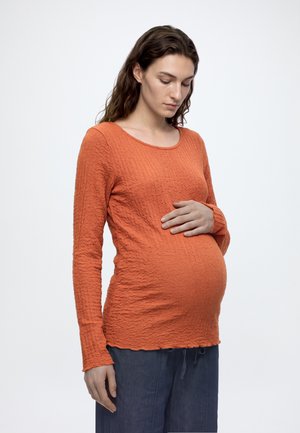 Mujer embarazada con cabello largo y castaño, vistiendo una camiseta naranja texturizada de manga larga y pantalones azules, sosteniendo suavemente su barriga.