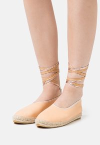 Espadrilhas em cor pêssego com sola de juta, apresentando tiras metálicas cruzadas ao redor dos tornozelos e um material superior liso.