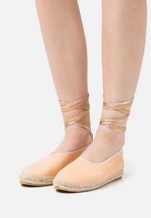 Espadrilhas em cor pêssego com sola de juta, apresentando tiras metálicas cruzadas ao redor dos tornozelos e um material superior liso.