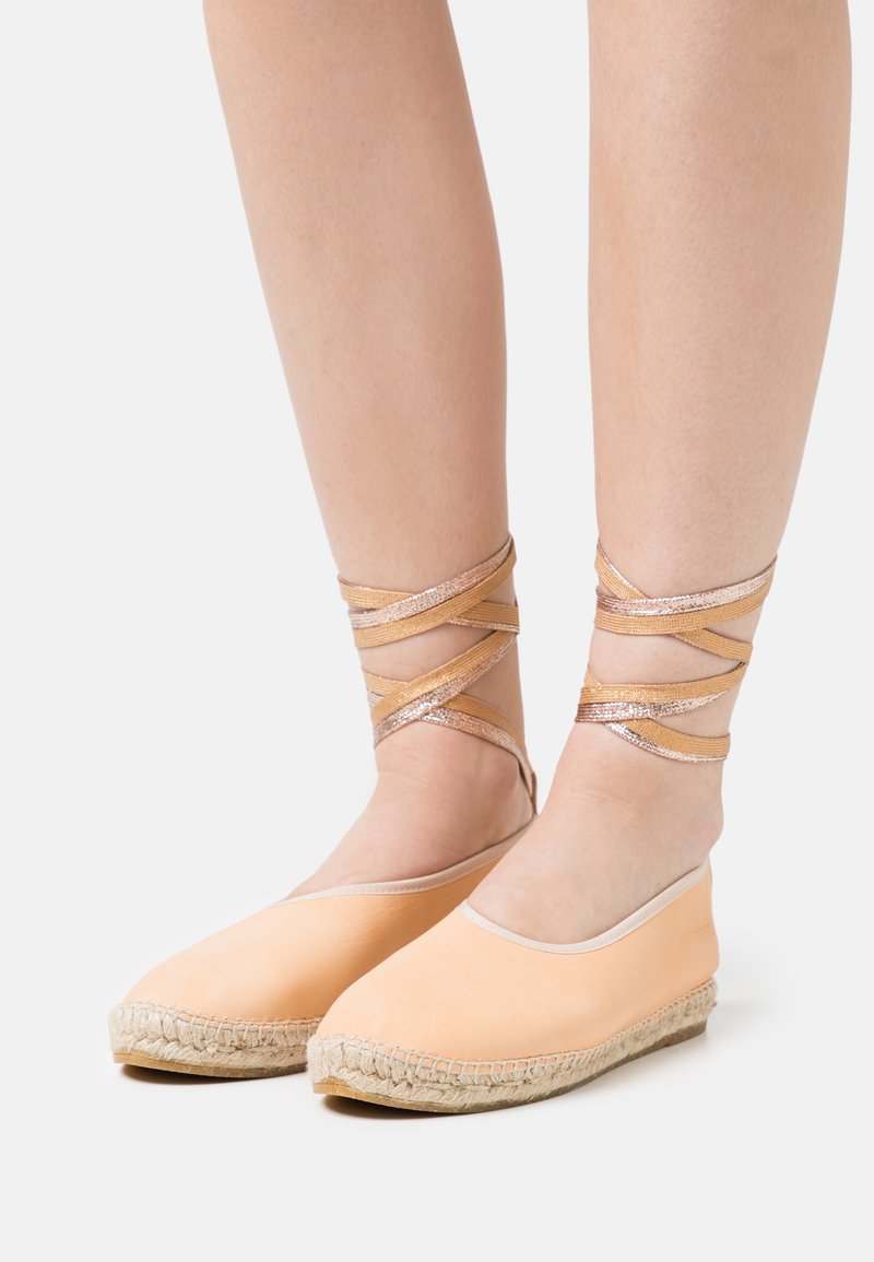 Espadrilhas em cor pêssego com sola de juta, apresentando tiras metálicas cruzadas ao redor dos tornozelos e um material superior liso.