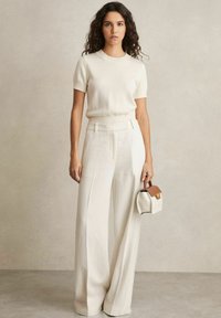 Maglione crema a maniche corte abbinato a pantaloni a vita alta e gamba larga. Porta una piccola borsa rigida con manico marrone.