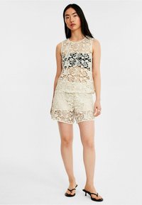 Lace sæt bestående af en ærmeløs top og shorts i lys beige med indviklede blomster mønstre og bølgede kanter. Sorte detaljer er synlige.