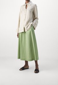 Camicia a righe crema e beige con collo a oblò, abbinata a una gonna midi verde plissettata e sandali neri in slide.