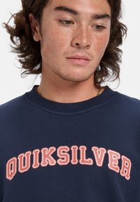 Námornícky sveter s krivým logom "QUIKSILVER" v tučnom koralovom písme. Kola krku, rebrované manžety a mäkká textúra látky.
