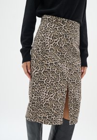 Pencil krilo s leopardjim vzorcem v bež in rjavi tkanini, ima sprednji razporek, visok pas in gladko teksturo. V kombinaciji s črnim zgornjim delom in škornji.