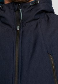 Uomo che indossa una giacca zip-up blu navy con cappuccio e cordini visibili vicino al collo.