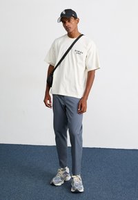 Cream short-sleeve T-shirt with logo, gray slim-fit pants, black cap, and multi-colored sneakers. Standing on blue carpet background.