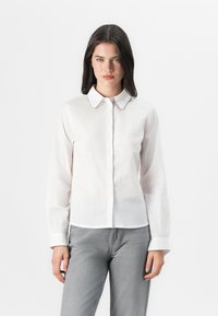 El modelo posa en una camisa blanca de botones con cuello y puños en contraste, combinada con jeans grises, frente a un fondo blanco liso.