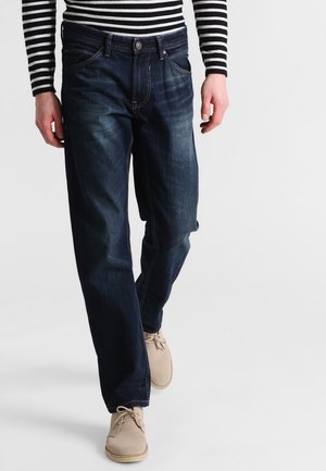 Dark blue denim jeans with a straight-leg fit, visible fading, and contrasting stitching. Paired with light beige shoes and a black-striped shirt.