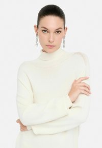 Pullover bianco a maglia con collo alto e polsini a coste, caratterizzato da un motivo testurizzato. La modella indossa orecchini argento a cerchio, con le braccia incrociate.